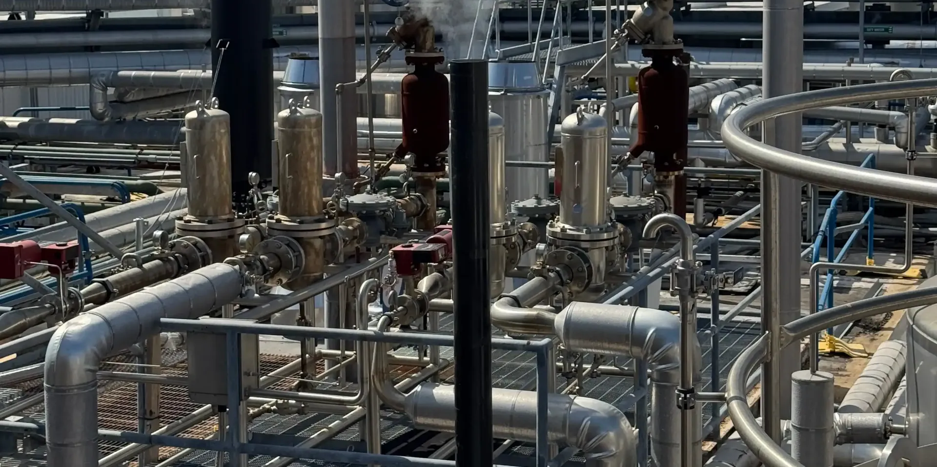 Industrial facility showing a complex network of metallic pipes, valves, and cylindrical pressure vessels.