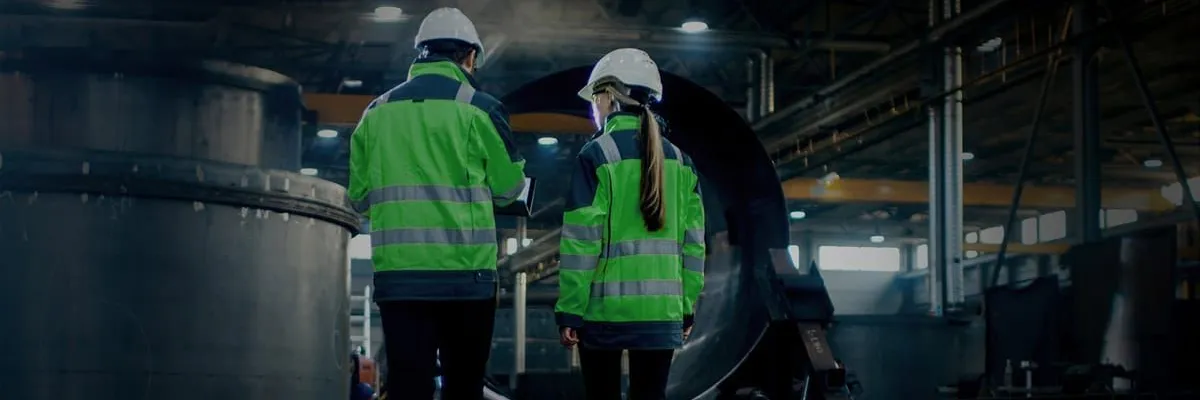 Two people in white hard hats and bright green safety jackets walk through a dimly lit industrial factory.